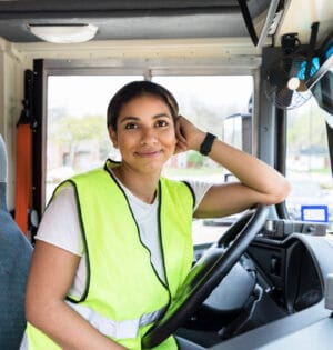 The cheerful, mid adult female bus driver leans on the steering wheel and smiles for the camera.